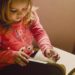 Little girl wearing a pink sweater with a teddybear on it, sitting reading a book.