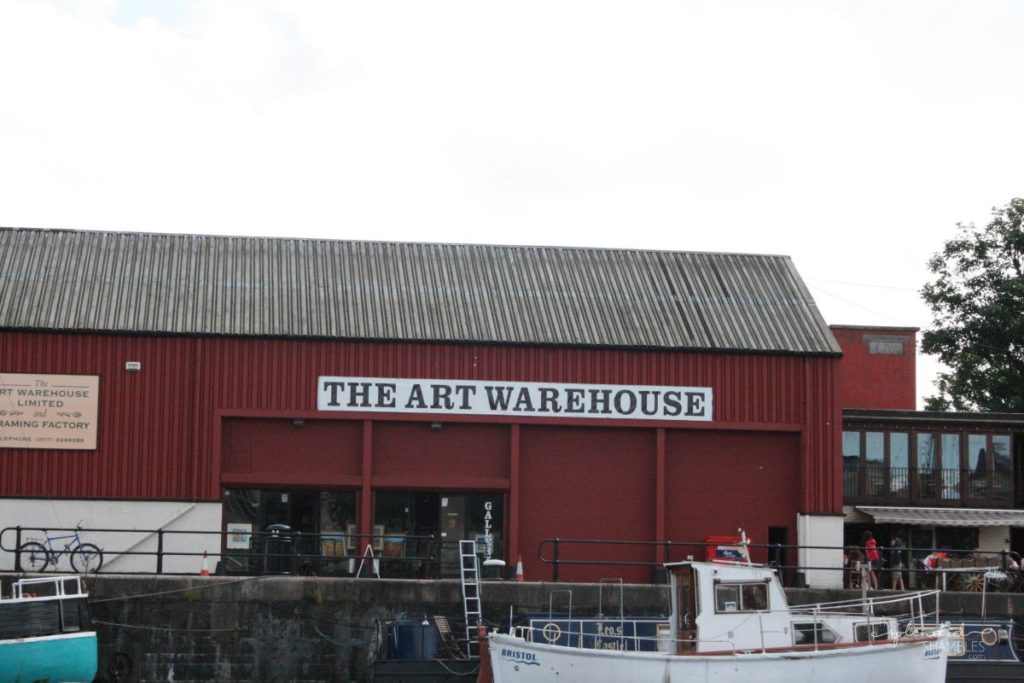 Red warehouse on the banks of the river, with the sign The Art Warehouse across the top. Boat in the foreground. How to spend a day in Bristol, England. This Splendid Shambles.