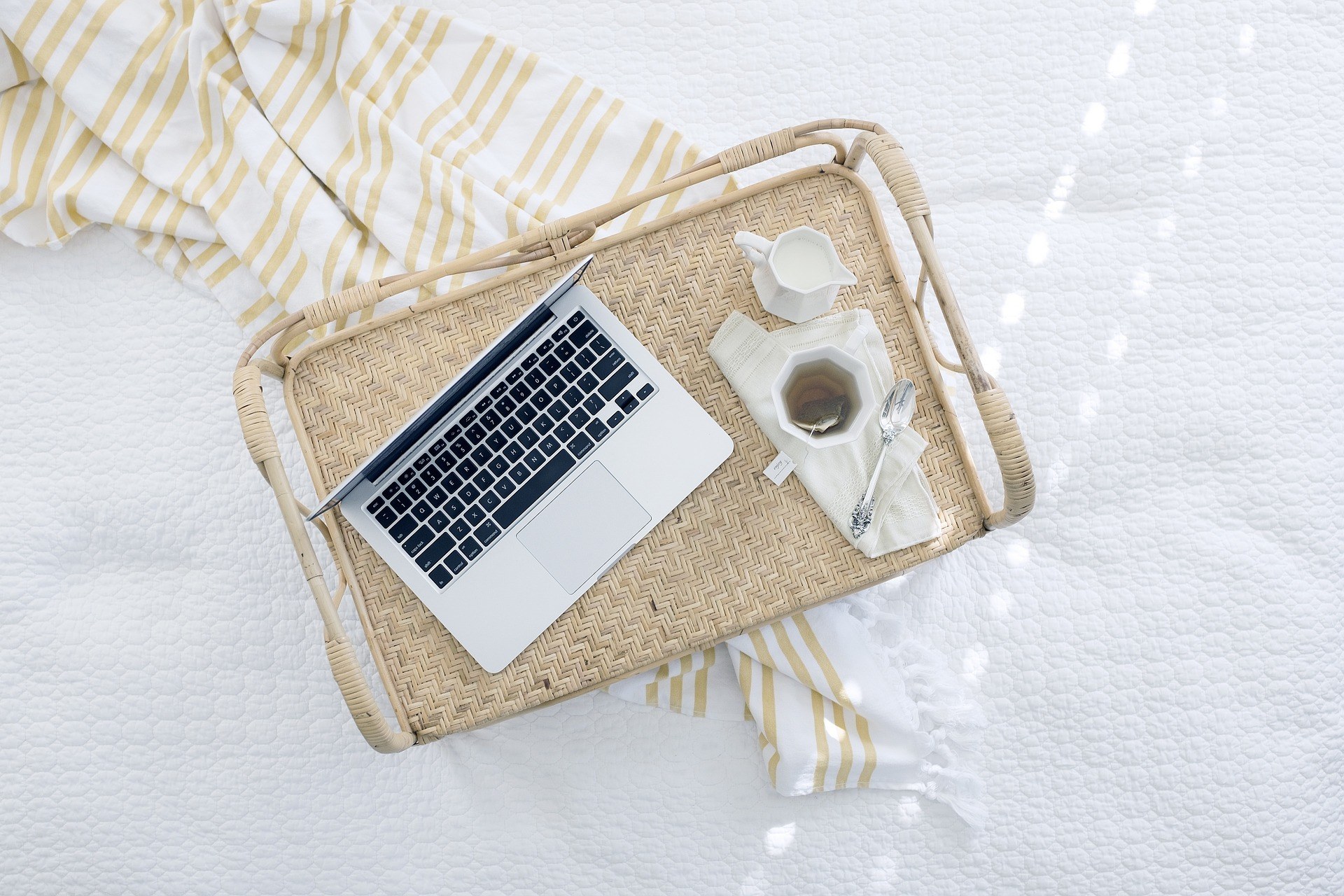 Apple laptop on a tray with a cup of tea. The tray sits on a bed with white covers and a yellow stripped blanket. What is social proof and why is it important? This Splendid Shambles