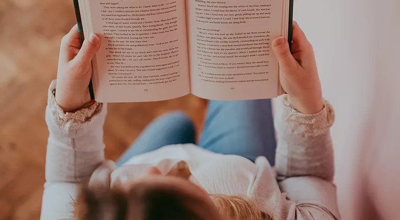 View from above, looking down on a woman reading an open book. The Bookish Mindset Interview.