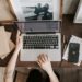 Close up of laptop on a dark wooden desk, arms of a woman around the laptop, one with fingers posed over the track pad. Mobile sitting on a notebook next to the laptop, and frames in the background. Blog post style errors you might be making, on This Splendid Shambles.