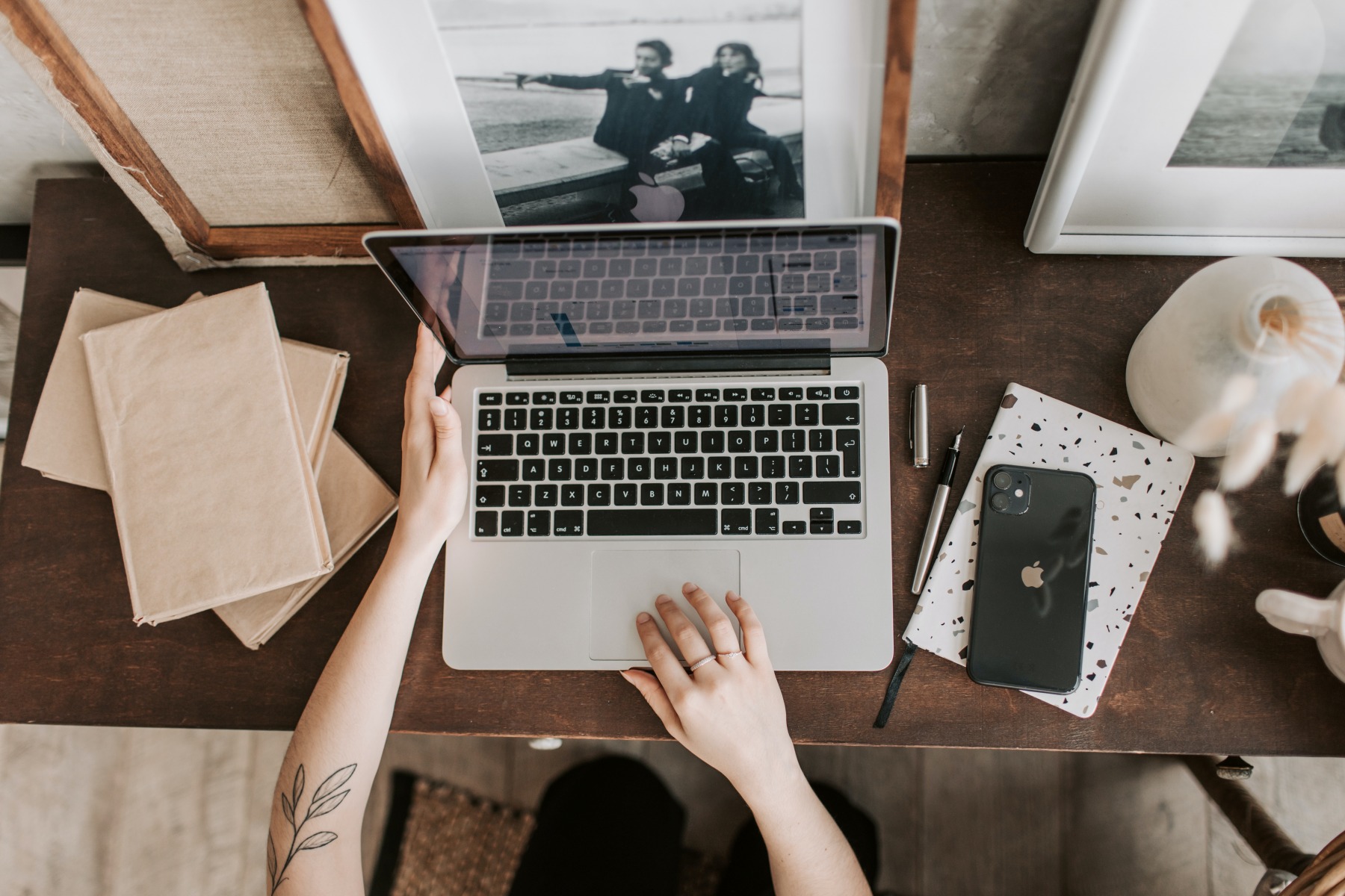 Close up of laptop on a dark wooden desk, arms of a woman around the laptop, one with fingers posed over the track pad. Mobile sitting on a notebook next to the laptop, and frames in the background. Blog post style errors you might be making, on This Splendid Shambles.