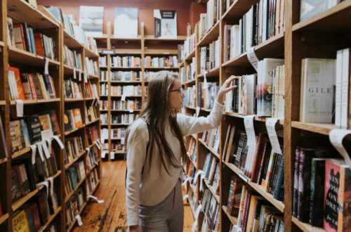 Woman in a bookstore, with her hand on a book, pulling it from the shelf. 8 Independent bookstores in New Zealand on This Splendid Shambles.
