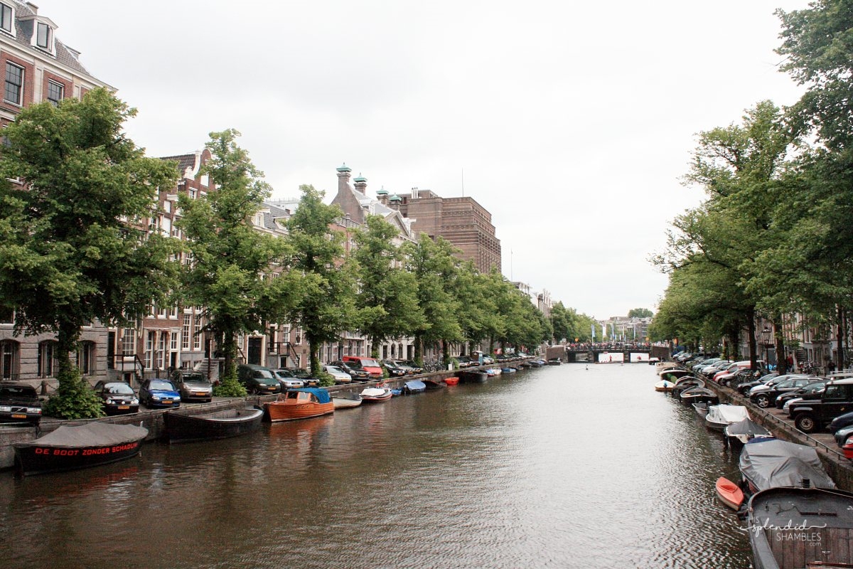 Amsterdam buildings on the edge of the canal, with boats in the water and tress along the edges. Pack Light for Amsterdam.