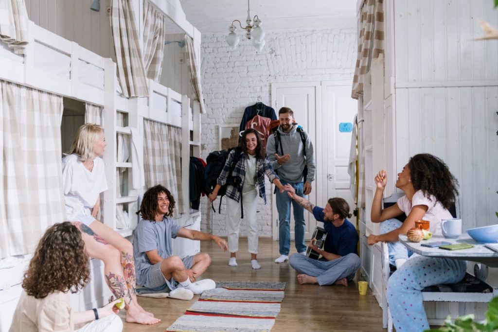 Two people walking into a room full of white bunk beds; five other people greeting them as they enter. Reasons to stay in a hostel while travelling. This Splendid Shambles. 