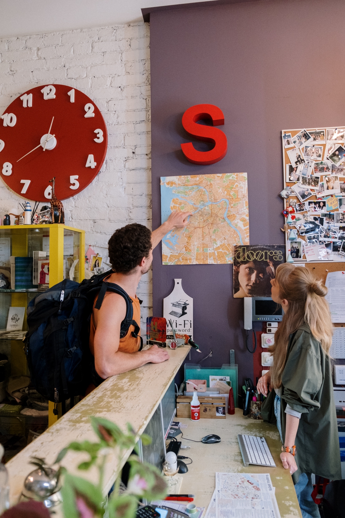 Man wearing a backpack pointing at map on the wall, over a reception desk. Woman at the desk also looking at the map. Reasons to stay in a hostel while travelling. This Splendid Shambles. 