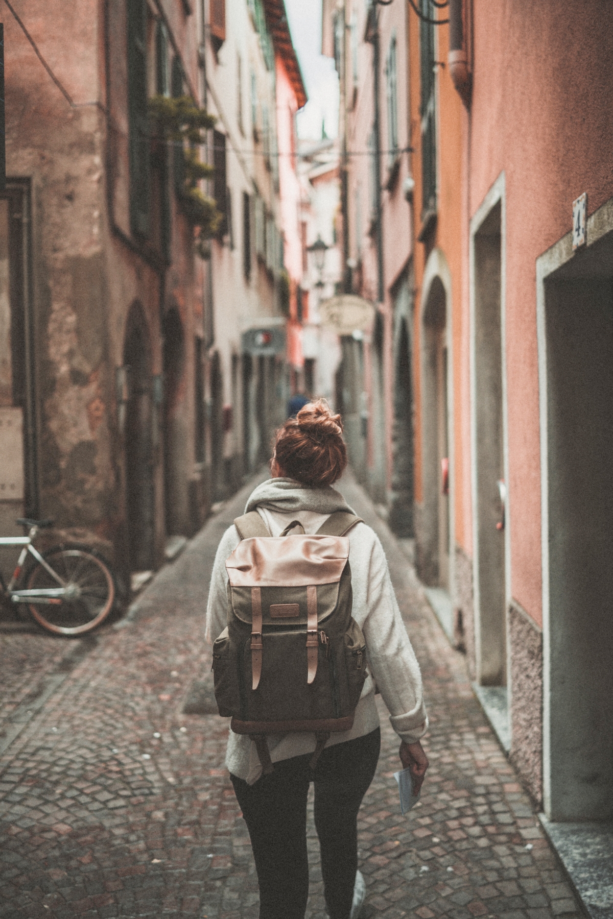 Woman facing away from the camera, facing towards an alley way in a European city, backpack on her pack. This Splendid Shambles Blog: Reasons why you should use a travel agent.