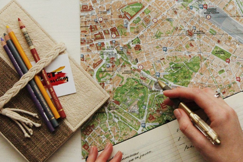 Map on a table, with coloured pencils to the left, and a hand holding a pen in the foreground. This Splendid Shambles Blog: Reasons why you should use a travel agent.