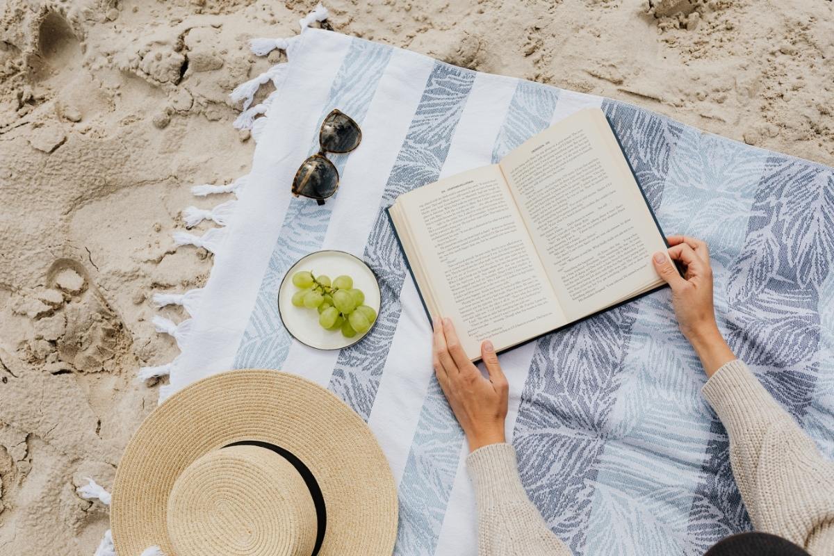 Picnic blanket on the beach, with summery items and arms holding a book.