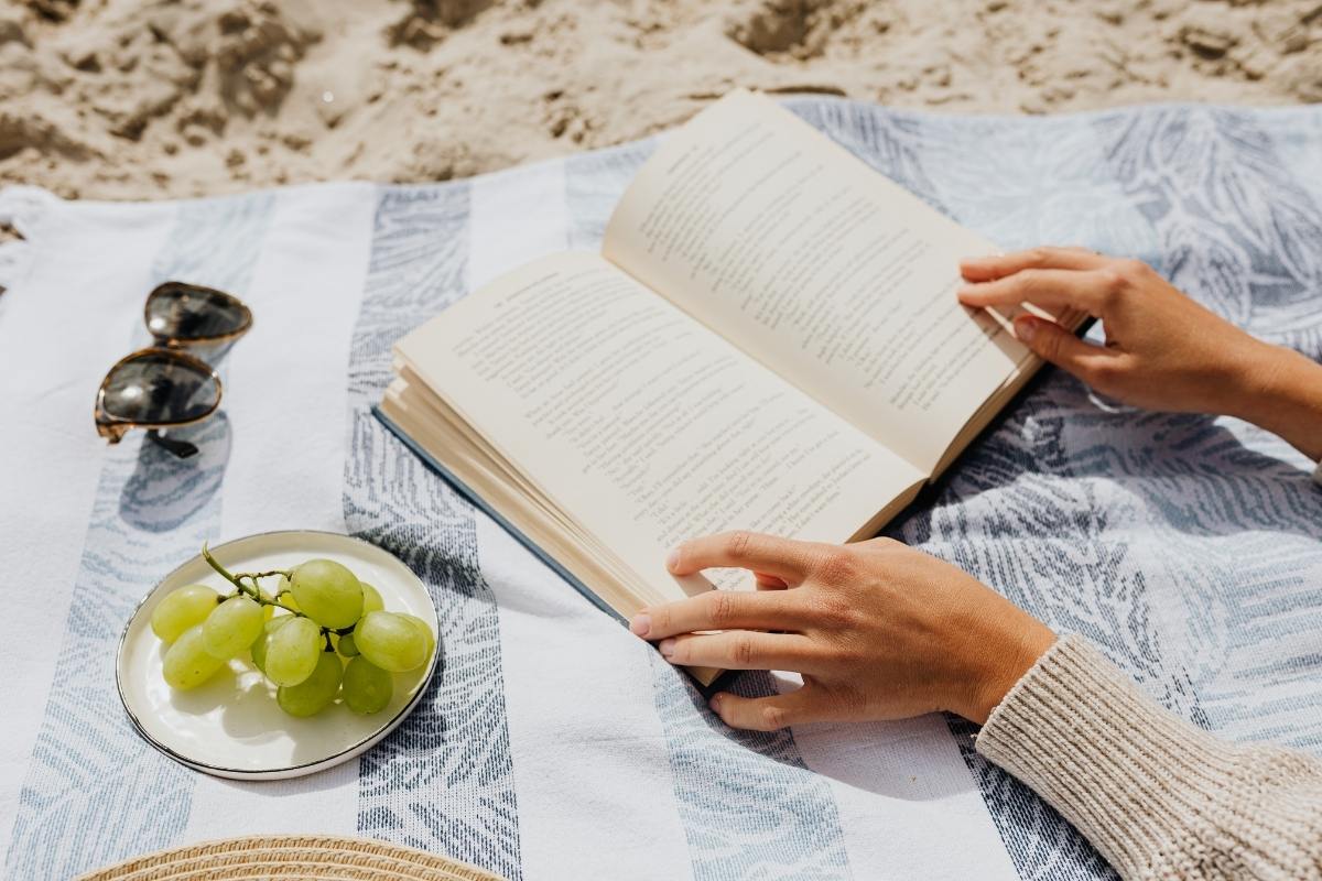Picnic blanket on the beach, with summery items and arms holding a book.