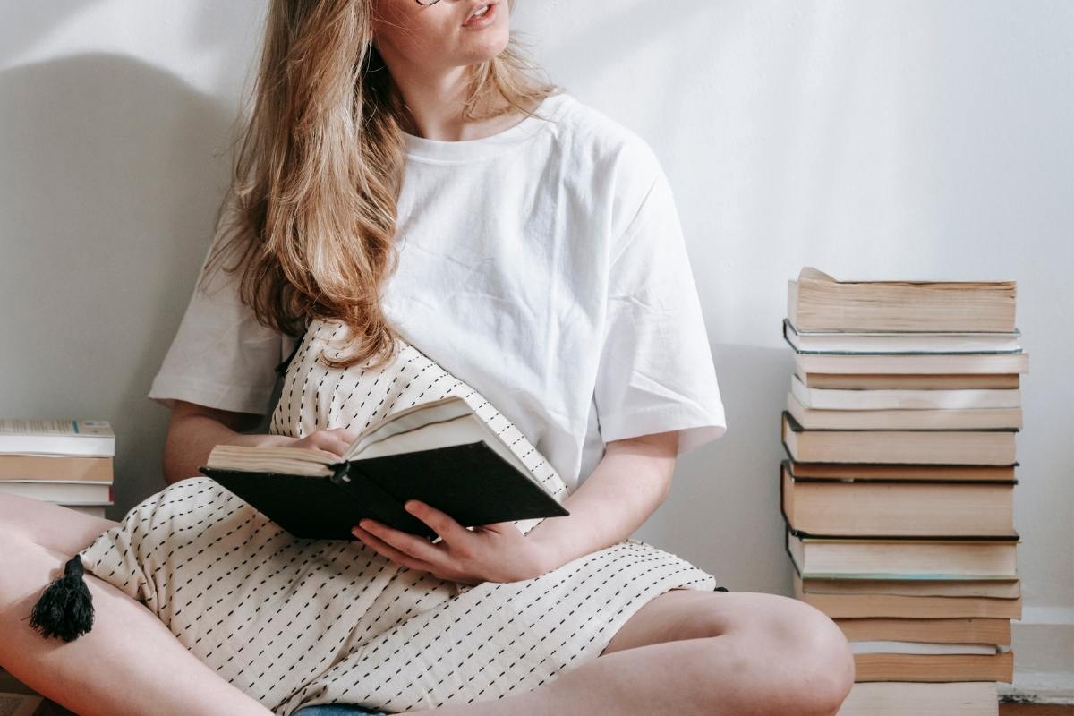 Woman sitting on the floor holding an open book. A stack of books is to her right. Young Adult Novel Series you need to read