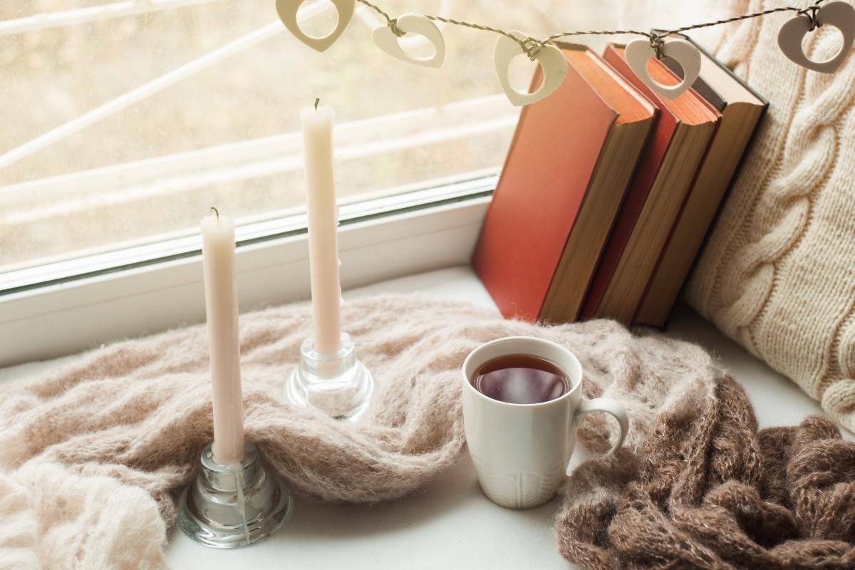 A stack of books propped up against a window, on a blanket. There is a cup of tea and two candles on the window sill as well.