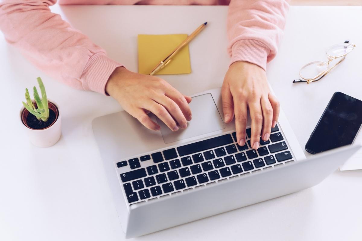 Woman in a pink sweater with her hands poised over a laptop. There's a yellow sticky note with a pen on top on the desk too. 5 Rules for writing your first guest blog post - This Splendid Shambles