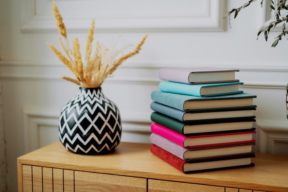 A pile of multi-coloured books sitting on a wooden sideboard, next to a blue and white vase with dried plants in it.