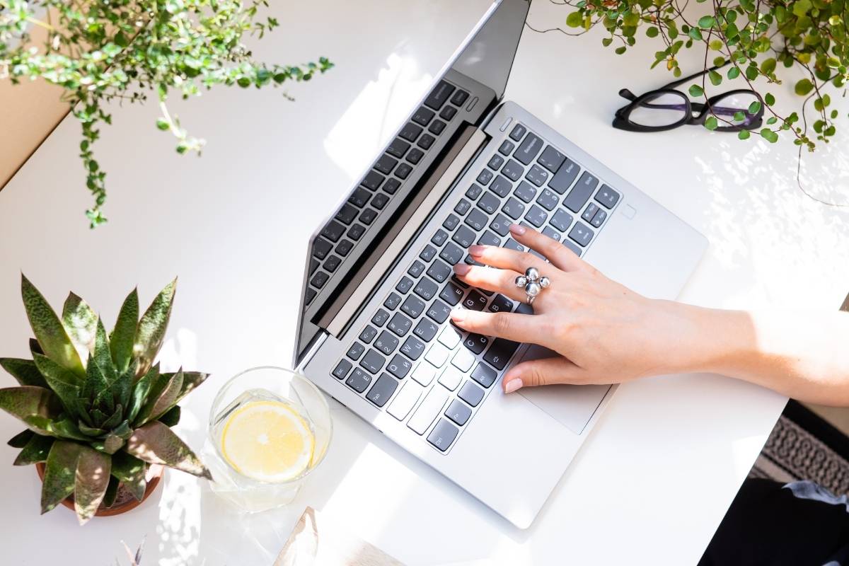 Woman's hand poised over a laptop, which is sitting on a white desk. There are plants around. Blogging For Small Business on This Splendid Shambles.