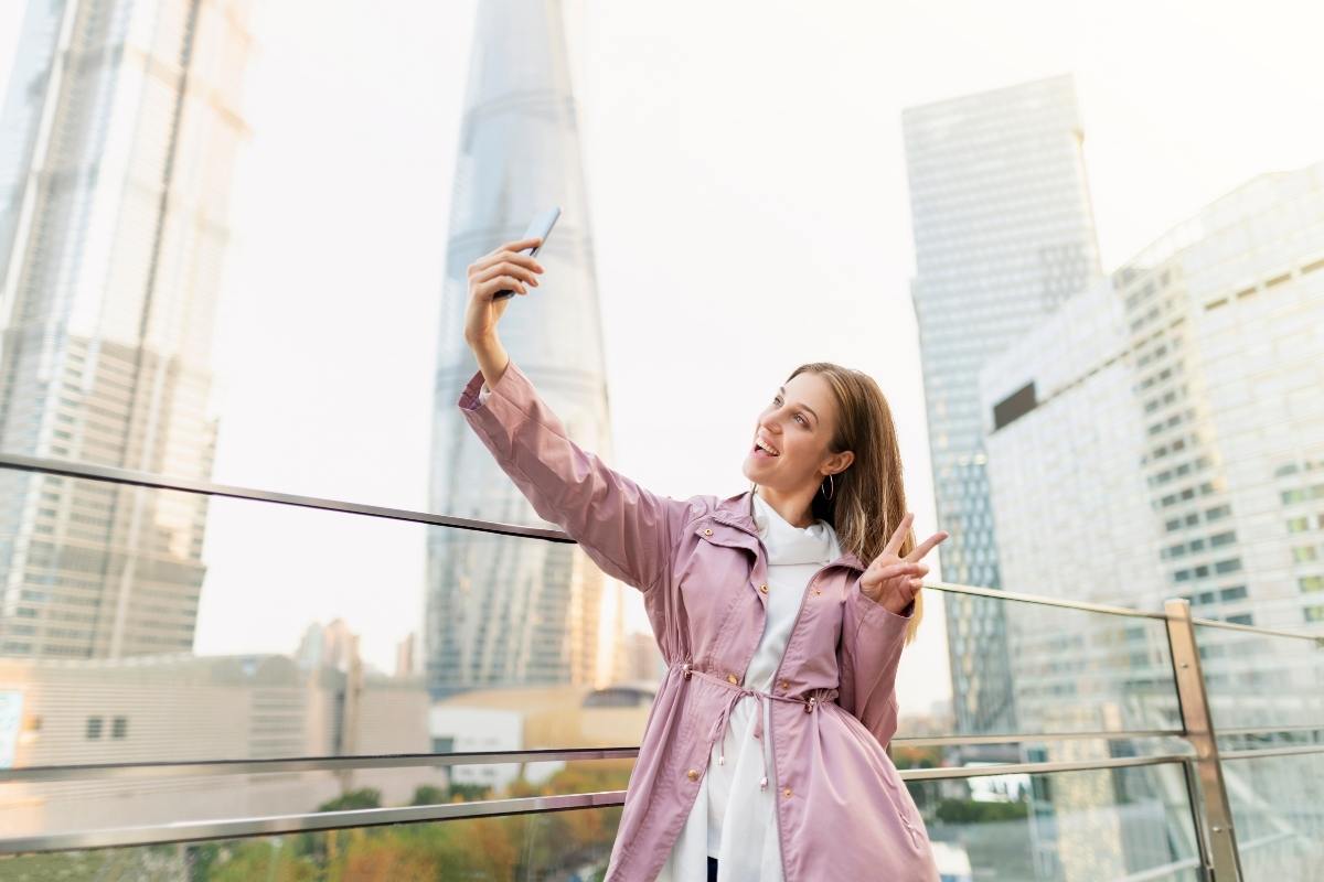 Woman in a pink jacket holding her phone up as if she's taking a photo or recording a video. She's also pulling the peace sign and smiling.
