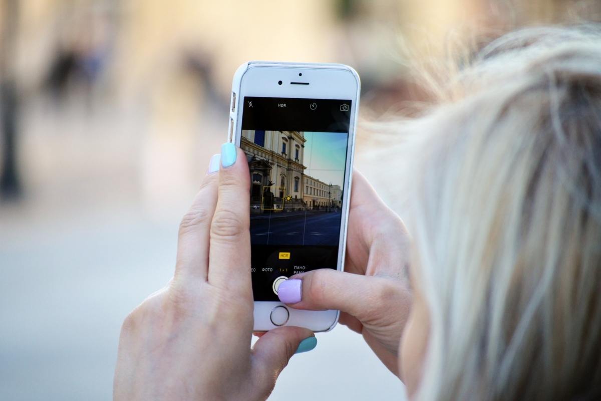 A woman with blonde hair facing away from the camera, holding up her smart phone and taking a photo. She has light pink names and the photo she is taking is of a building.