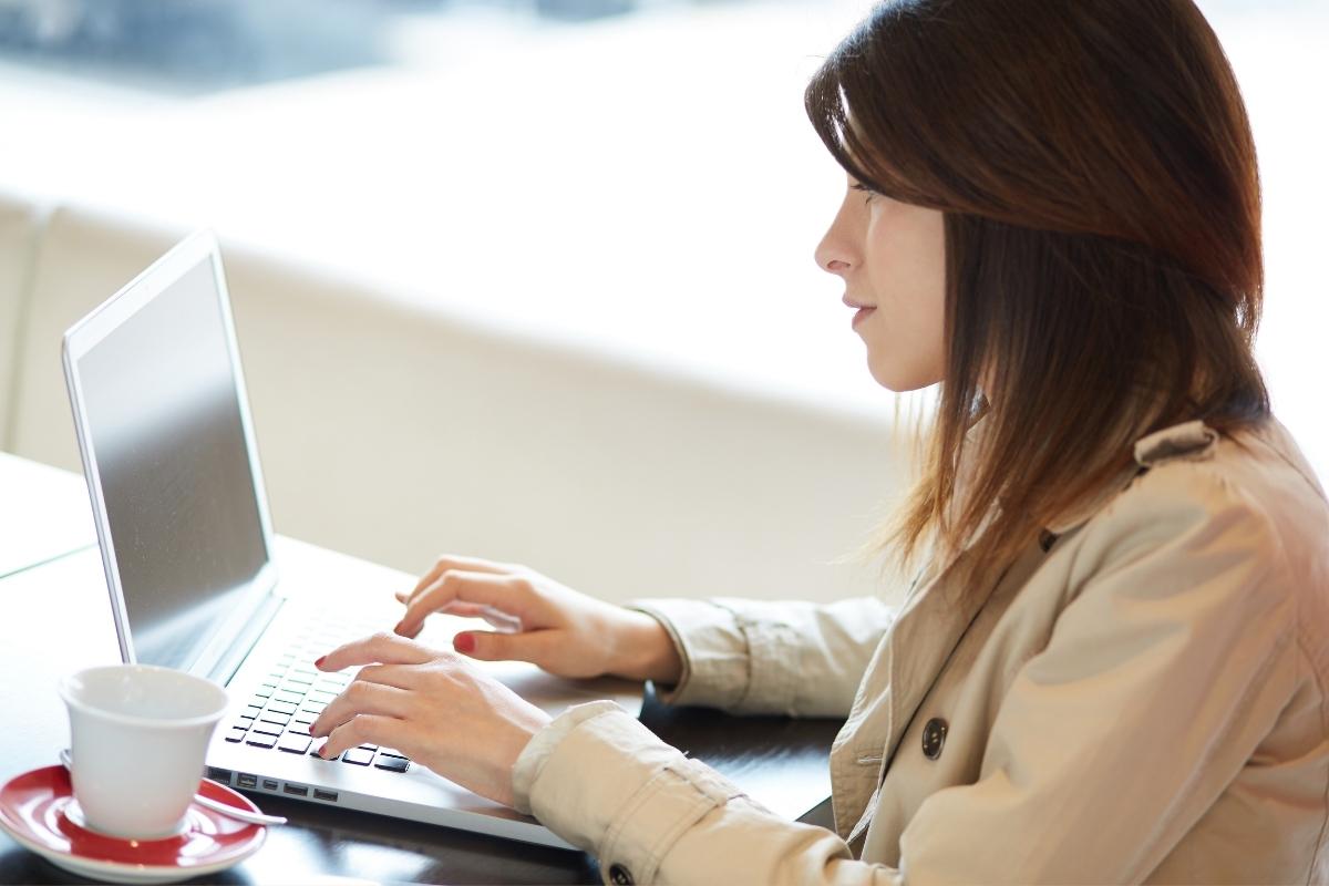 Side profile of a woman with dark hair sitting at a table, fingers on her lpatop keyboard, with a white coffee cup to her left. 