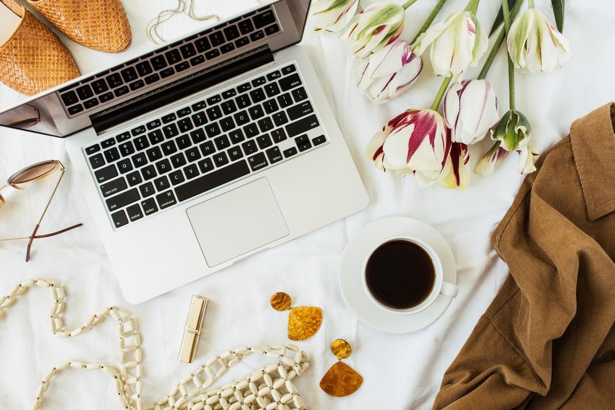 Flat lay of a white surface, with laptop, coffee mug, flowers, earrings and other lifestyle items around the edges.