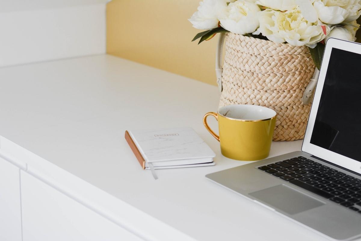 Laptop to the right of the screen, sitting on a desk. There is a basket of flowers and a yellow mug alongside a notebook.