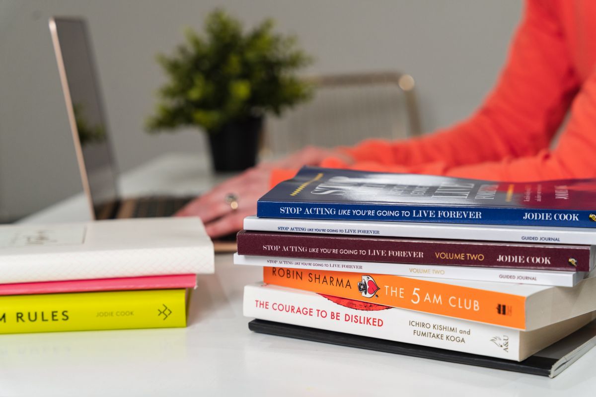 Woman in an orange in the background, fingers poised over a laptop. In the foreground there are two piles of non-fiction books. 