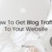 Apple laptop, a coffee and a sweet treat on a white blanketed bed, with the words 'how to get blog traffic to your website' over the top.