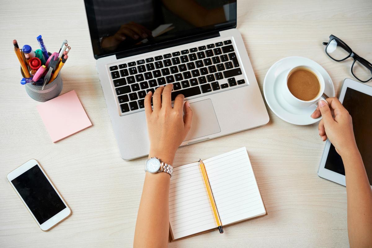 Hand poised over a laptop keyword, cup of tea to the right, notebook to the bottom and other stationery to the left. 