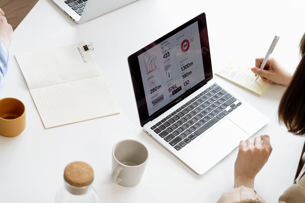 Open laptop on a white desk, coffee to the left. There are business stats on the laptop screen. 
