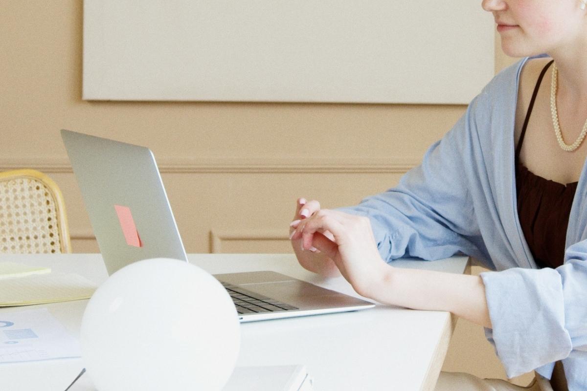 Woman in a blueshirt leaning on a white table with laptop open. 