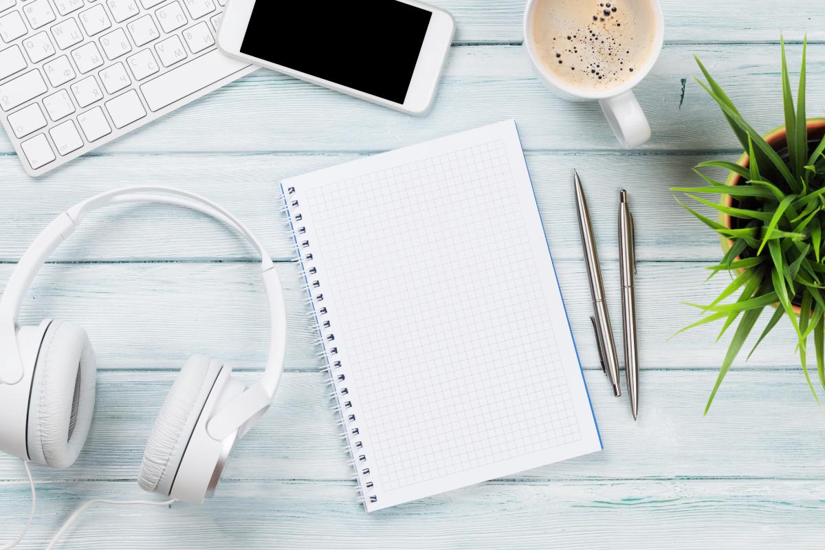 Notebook, pens, phone, headphones and the corner of a coffee cup on a blue-wooden desk/table.